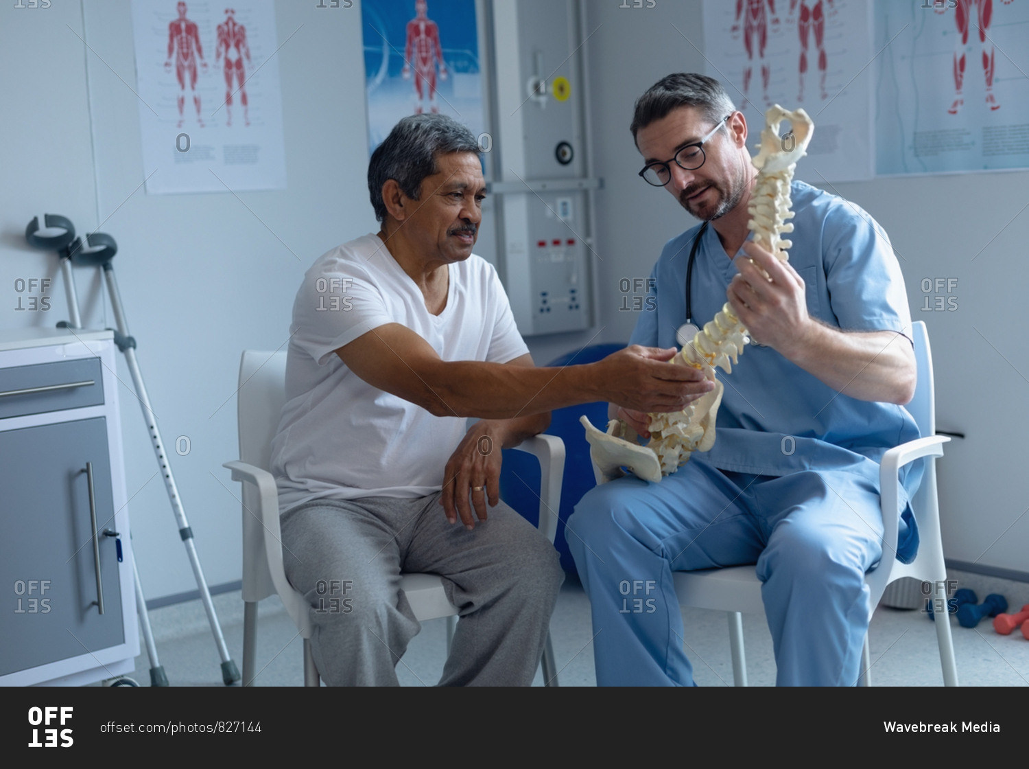 Front view of handsome Caucasian male orthopedic surgeon explaining spine model to mixed-race male patient in hospital