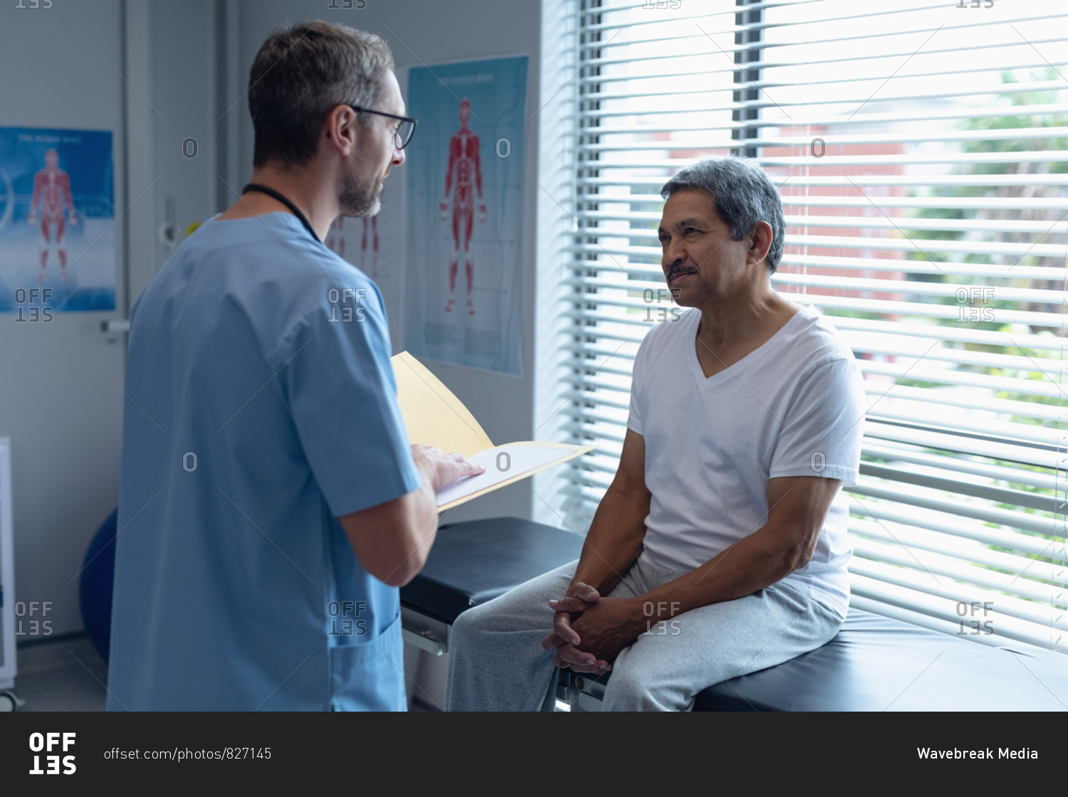 Side view of handsome Caucasian male surgeon interacting with senior mixed-race patient while holding medical files in his hands in hospital