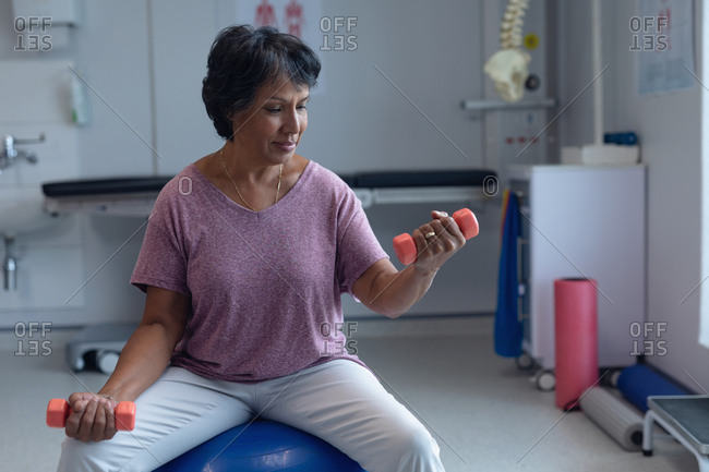 Front view of beautiful mixed-race female patient exercising with dumbbells on exercise ball  in the hospital