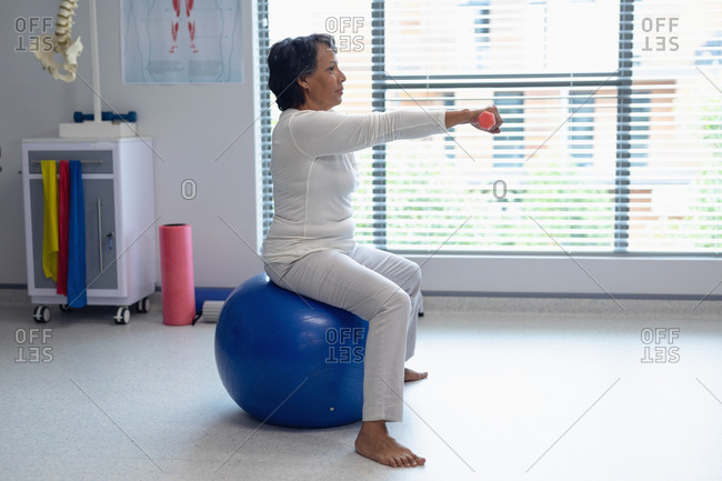 Side view of mixed-race female patient exercising with dumbbells on exercise ball in the hospital
