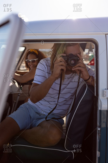 Front view of young Caucasian man taking pictures with digital camera in front seat of camper van at beach. Mixed race woman smiling in the background