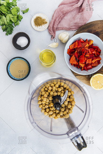 Cooked chickpeas in a blender, chopped roasted red peppers, tahini, olive oil and other hummus ingredients photographed on a grey background