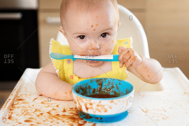 Funny messy baby in high chair with spoon in mouth