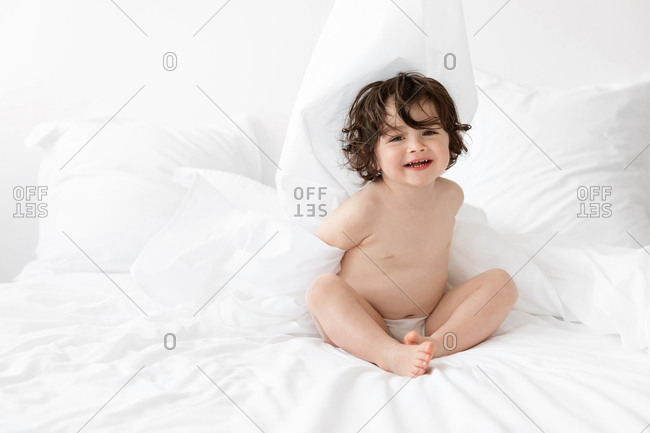 Playful young child sitting on white bed smiling