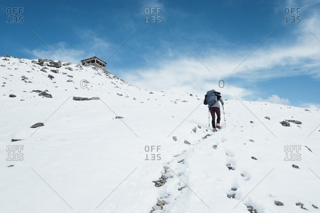 Female hiker treks upwards through snow to abandoned building at mountain peak