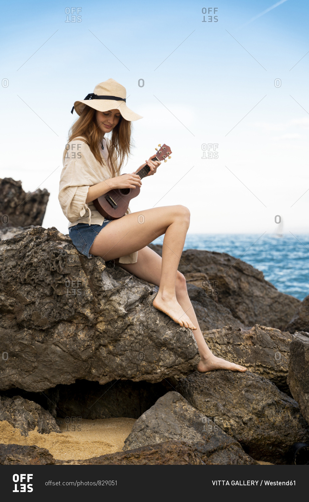 Smiling young woman wearing summer hat sitting on rock playing ukulele
