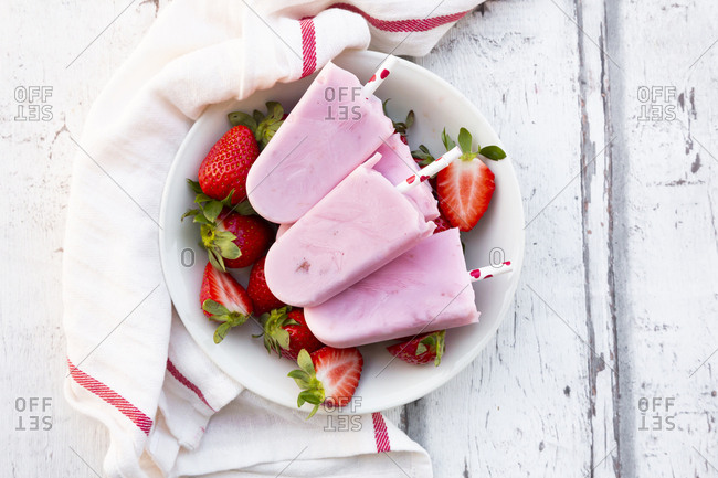 Homemade strawberry yogurt ice lollies with fresh strawberries in a bowl on white wood
