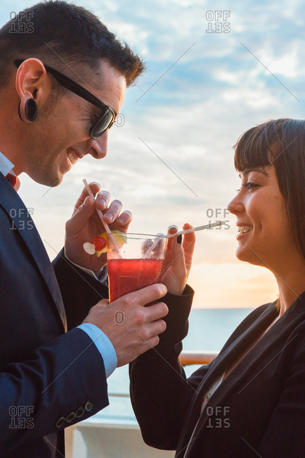 Side view of young attractive couple drinking red beverage with straws from one glass on background of sunset sea