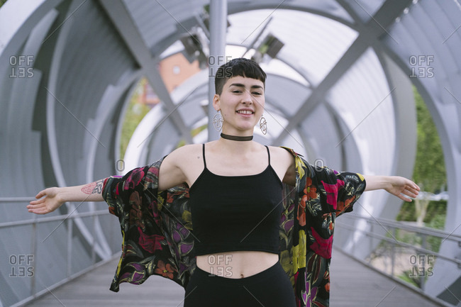 Young brunette woman in colorful shirt and black clothes standing with hands spread wide in round tunnel and looking at camera