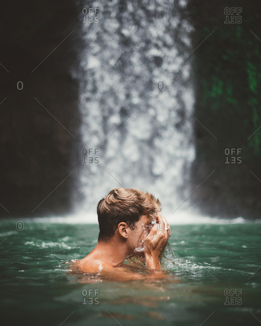 Side view of man washing face while swimming in clear water of lake with waterfall on background, Bali
