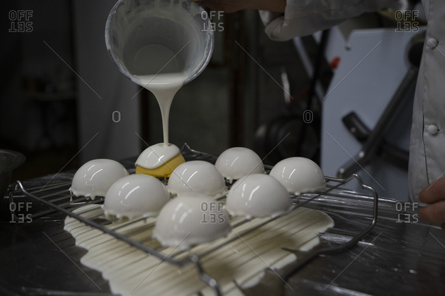 Crop person pouring white mirror glaze on tasty yellow mousse cakes arranged on metal grid in kitchen