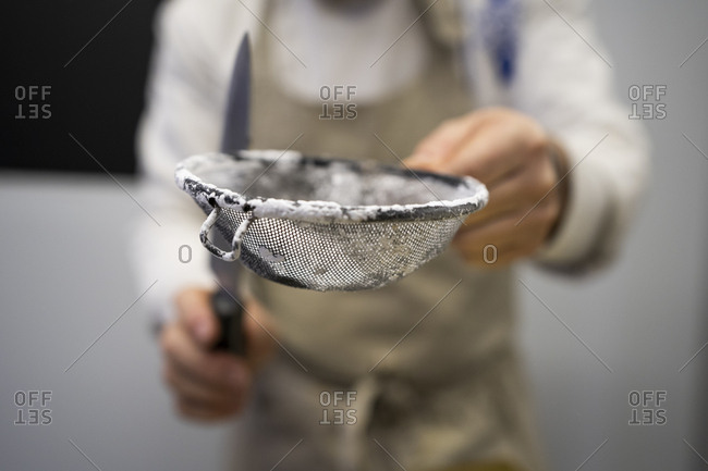 Crop man in apron standing and holding metal sieve with flour and knife on blurred background
