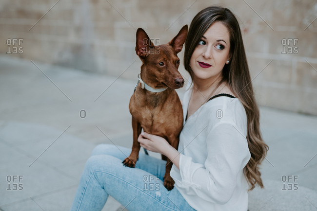 Beautiful casual woman with hound dog sitting on concrete step on street looking away