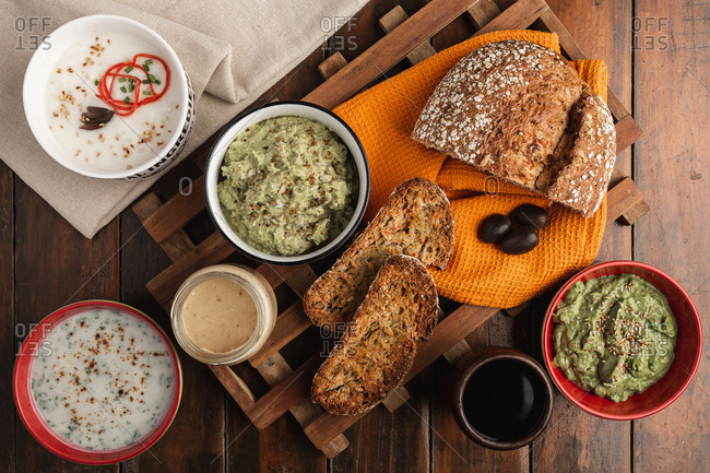 From above appetizing different sauces in red white bowls with sesame seeds toppings and sliced fragrant toasted freshly baked bread on wooden surface