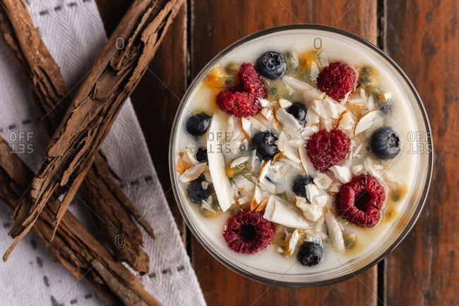 From above appetizing fragrant yogurt with raspberry currant oat in glass and red bowls decorated with cinnamon sticks on wooden background