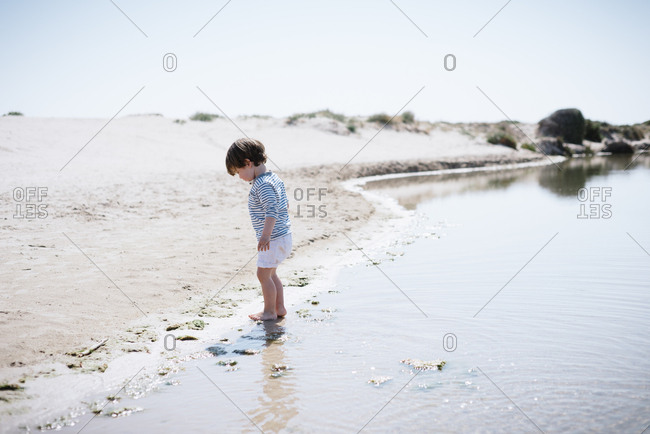 Side view of cute baby standing with bare feet in sand and looking at shiny calm sea in beautiful beach in sunny day