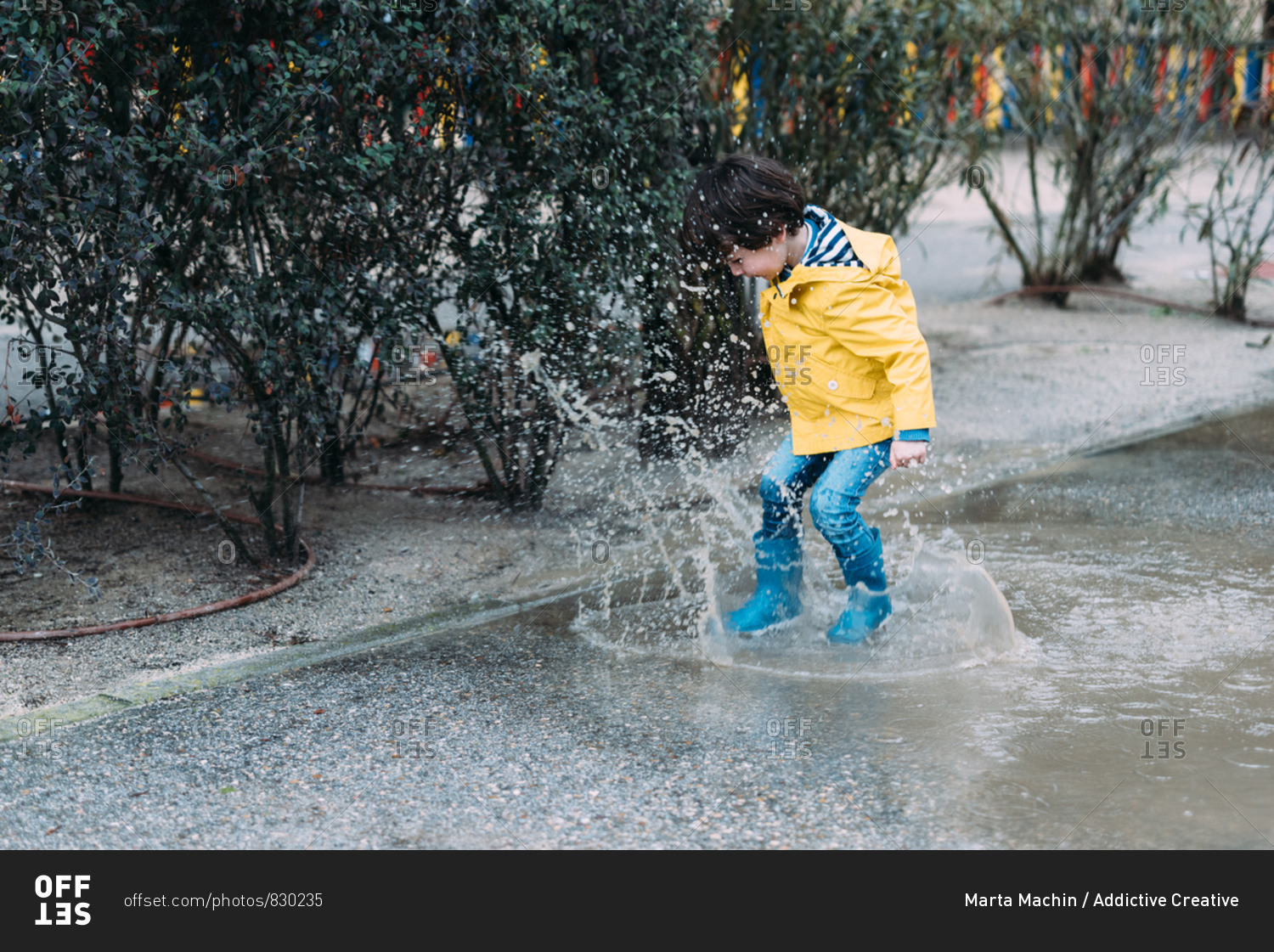 Excited boy in coat and gumboots having fun on street and jumping on puddle