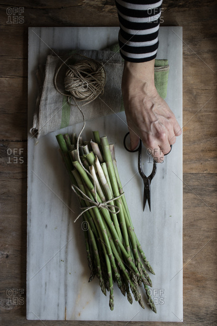 Top view of person hands holding scissors in marble board with pile of asparagus tied with twine rope on wooden table