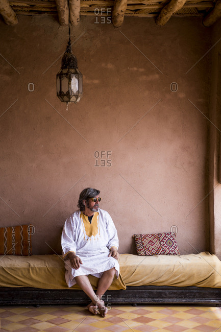 Adult man in long clothes sitting on sofa on terrace with stone fence in oriental style, Morocco