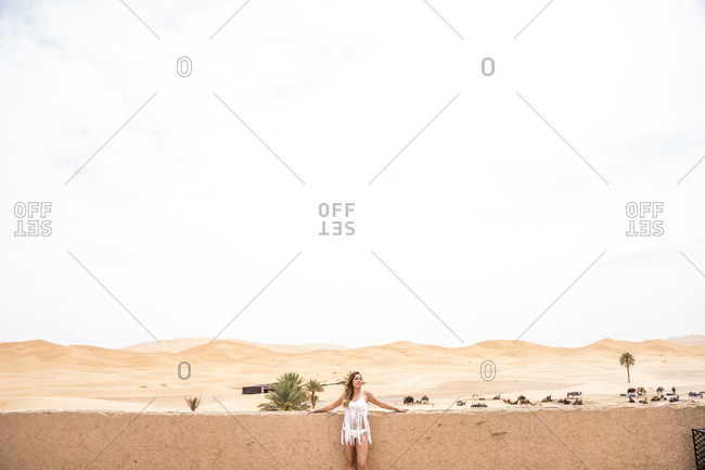 Beautiful young woman in white top leaning on a wall looking away against endless sandy desert, Morocco