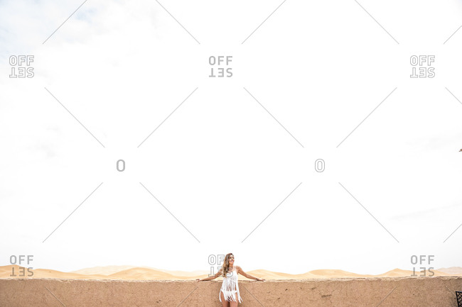 Beautiful young woman in white top leaning on a wall looking away against endless sandy desert, Morocco