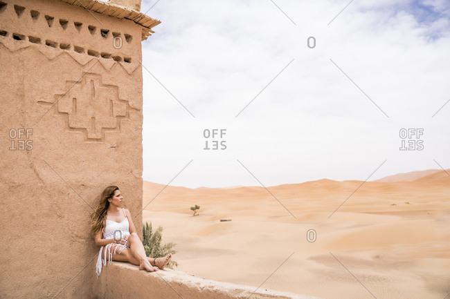 Beautiful young woman in white top sitting on stone fence in wind looking away against endless sandy desert, Morocco