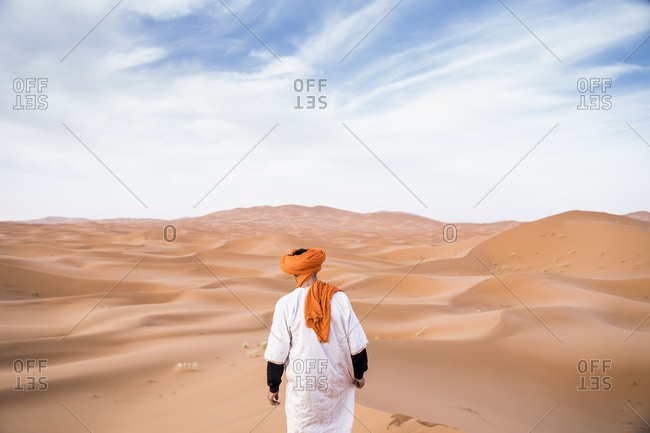 Back view of man wearing long outfit with colorful turban walking on dunes of endless sandy desert, Morocco
