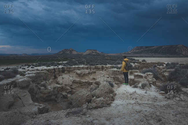 Side view of young man in yellow jacket and backpack standing on stone and looking at impressive blue sky in twilight in semi-desert Bardenas Reales Navarra Spain