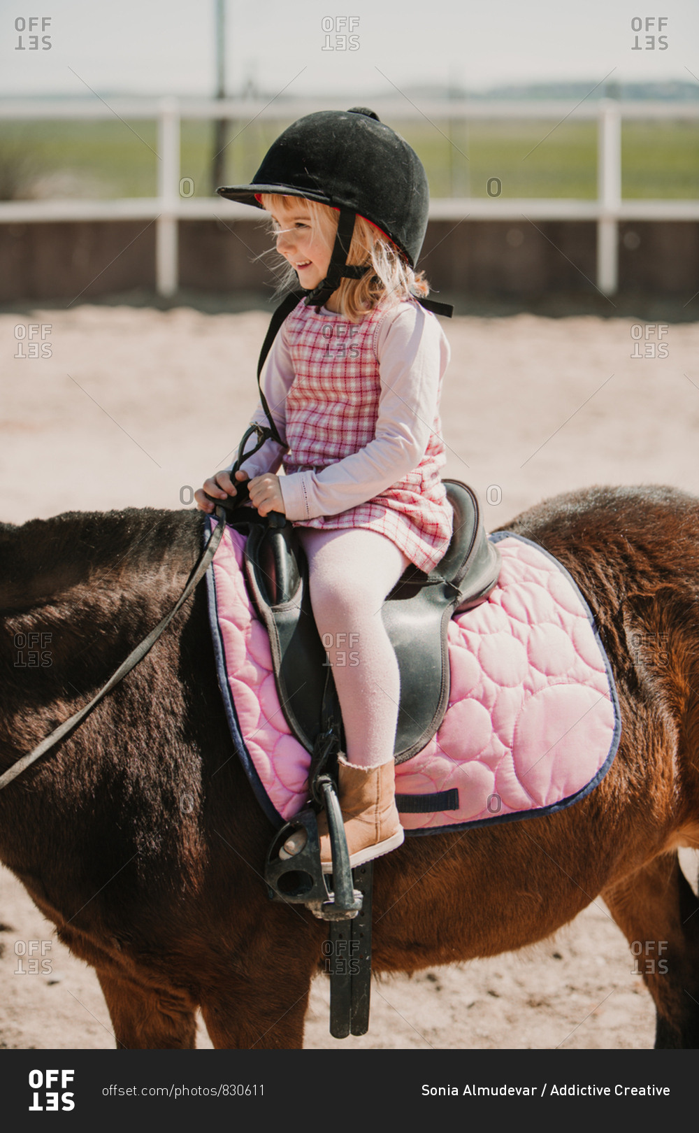 Cheerful small girl in dress and jockey hay sitting on horse while learning to ride on racetrack