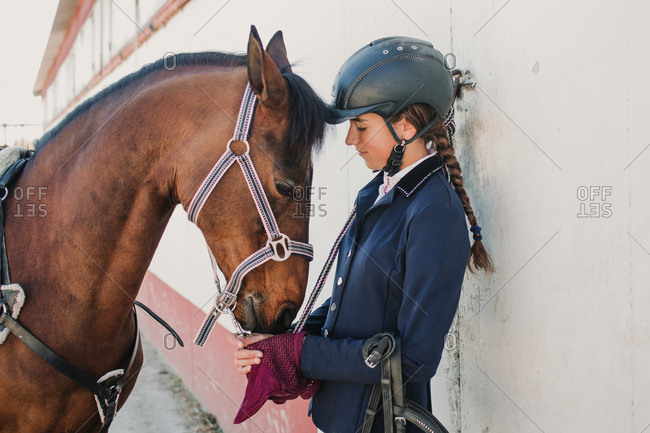 Side view of young teen woman in jockey helmet and jacket caressing horse standing together outdoors