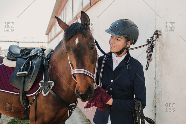 Side view of young teen woman in jockey helmet and jacket caressing horse standing together outdoors