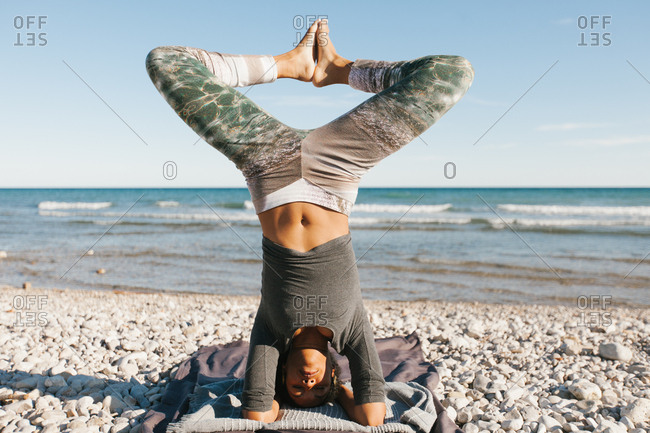 African American attractive young woman in yoga headstand posture in open air on background of calm water in sunny day