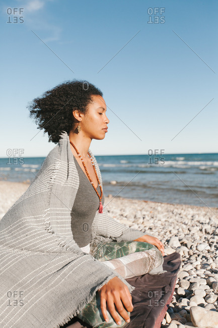 Side view of African American attractive young woman covered with grey plaid meditating in lotus yoga posture on sandy beach in bright day
