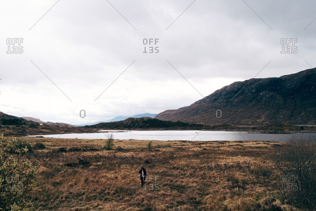 Adult man with backpack standing in picturesque remote valley with mountains and lake looking away