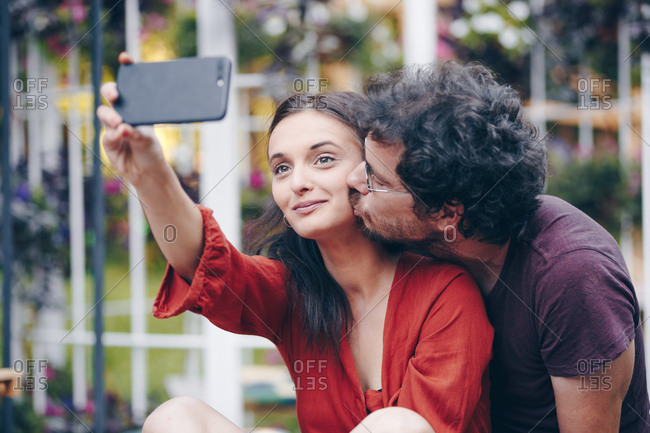 Authentic image of beautiful young couple in love taking selfie on smartphone during summer music festival. Boyfriend giving his girlfriend a kiss.