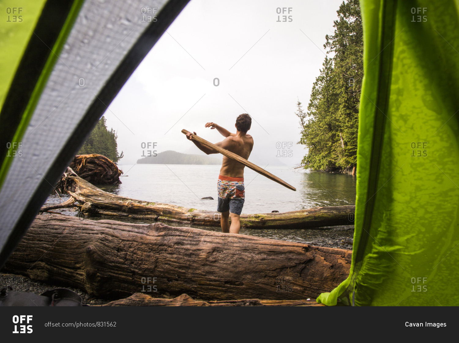 KLEMTU, BRITISH COLUMBIA, CANADA. A shirtless man swings a large stick like a baseball bat on a remote beach, as seen through the opening in a green tent door.