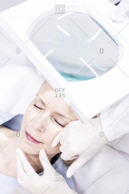 Beauty technician examining woman's facial skin using magnifying glass, close-up.