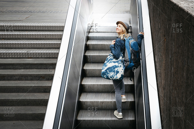 Woman with backpack and travelling bag standing on escalator looking up
