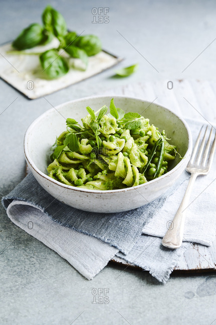 A garden pea pesto pasta in a bowl with fresh peas and seen from a slight angle with a fork on the side and basil in the background.