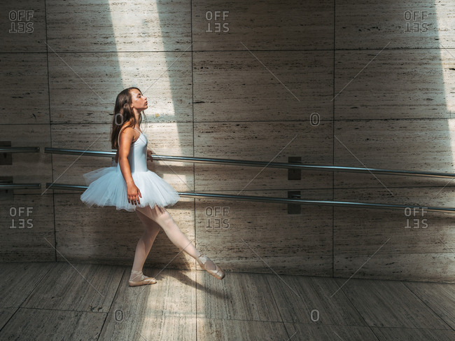 Ballerina in white ballet tutu performing exercises at rail in contrast sunny light