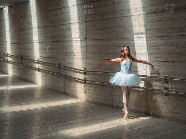 Ballerina in white ballet tutu performing exercises at rail in contrast sunny light