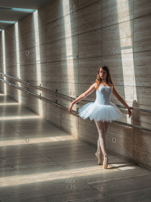 Ballerina in white ballet tutu performing exercises at rail in contrast sunny light