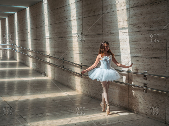 Ballerina in white ballet tutu performing exercises at rail in contrast sunny light