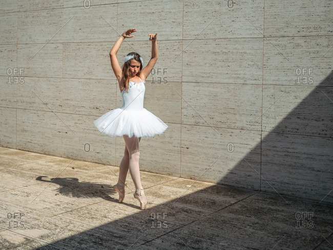 Ballerina in white ballet tutu performing exercises in classroom in contrast sunny light