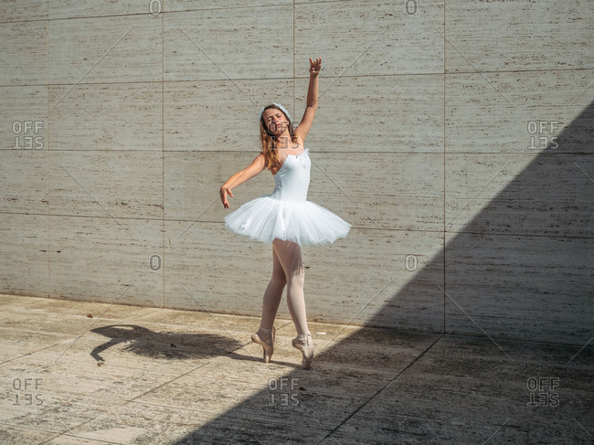 Ballerina in white ballet tutu performing exercises in classroom in contrast sunny light