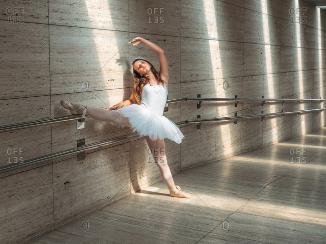 Ballerina in white ballet tutu performing exercises at rail in contrast sunny light