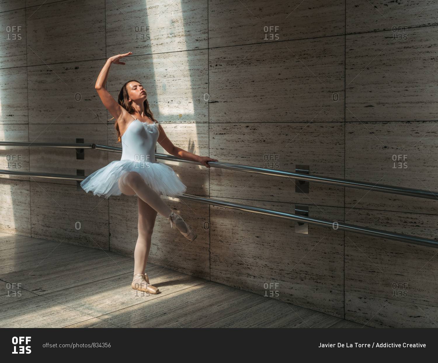 Ballerina in white ballet tutu performing exercises at rail in contrast sunny light