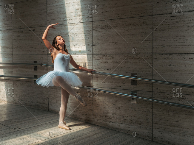 Ballerina in white ballet tutu performing exercises at rail in contrast sunny light