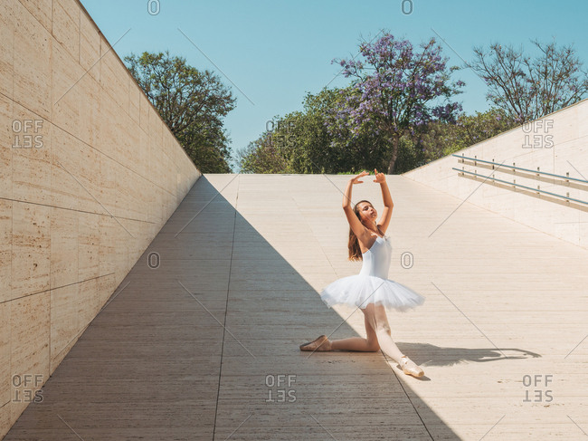 Classical ballet position gracefully performing by ballerina with raising hands and stretching legs outside in bright sunny day