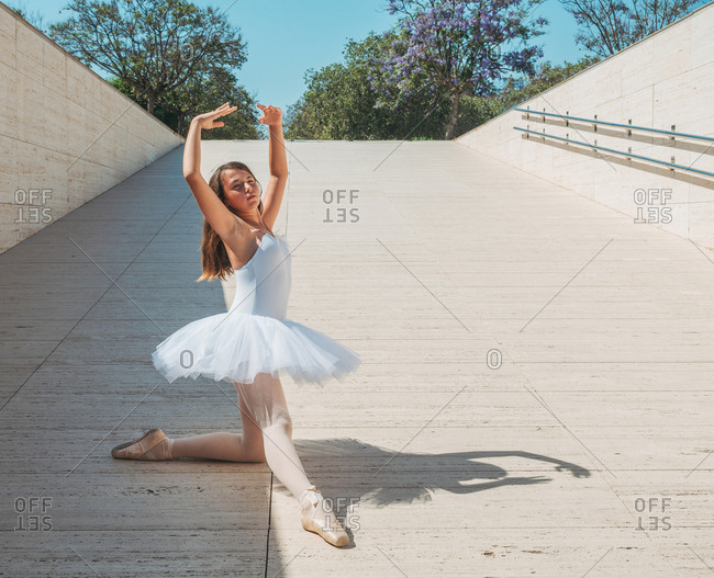 Classical ballet position gracefully performing by ballerina with raising hands and stretching legs outside in bright sunny day